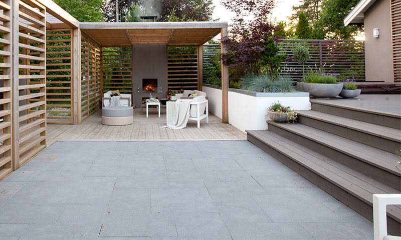 Lyst sitteområde under pergola med peis, grå fliser, tretrapp og grønne planter