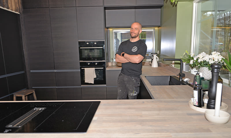 Moderne kjøkken med mørke skap, lys trebenk, integrert kokeøy, hvite blomster ved vindu og person