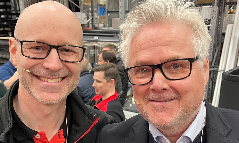 To menn i svarte og røde jakker står i travelt varehus med høye metallreoler.