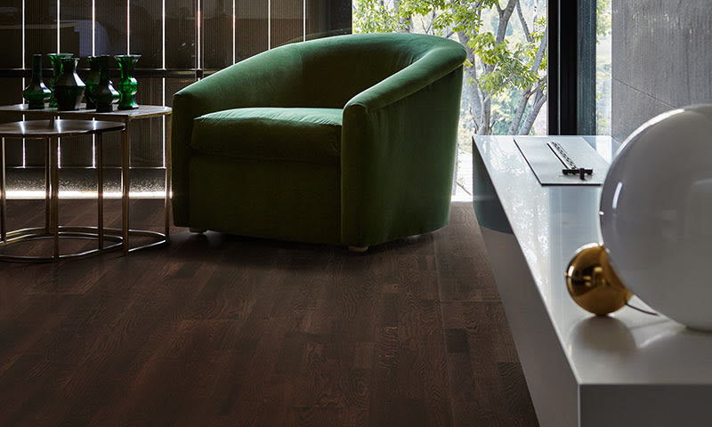 Grønn fløyelslenestol ved vindu med utsikt til trær, mørkt tregulv og små bord.