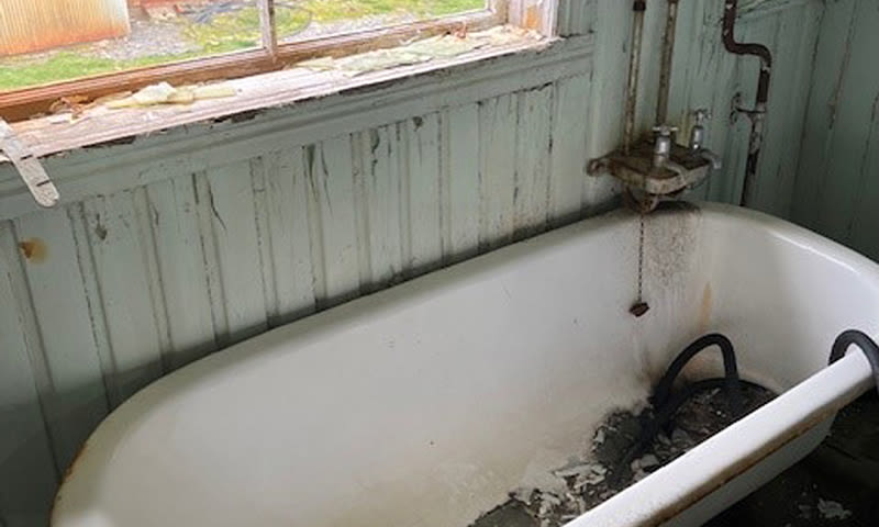 Falmet grønn panelvegg bak slitt hvitt badekar med rust, smuss, knust vindu og liten vask.