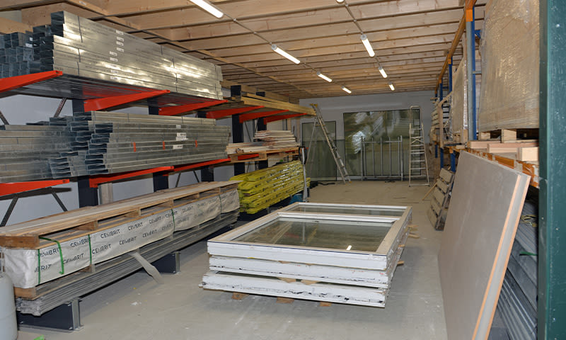 Byggevareslager med røde stålhyller, gule isolasjonspakker og stabler av hvite vindusrammer.