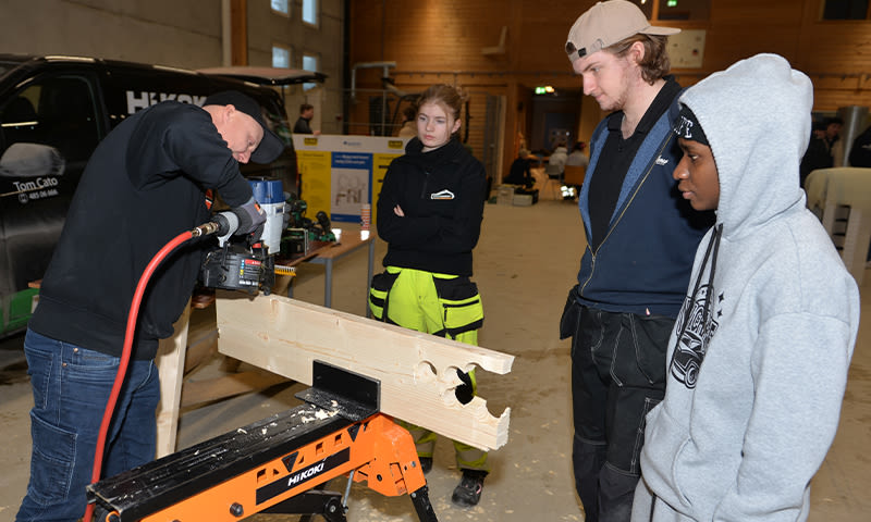 Mann bruker spikerpistol på trestokk festet i oransje HiKOKI-stillas mens tre i arbeidsklær ser på.