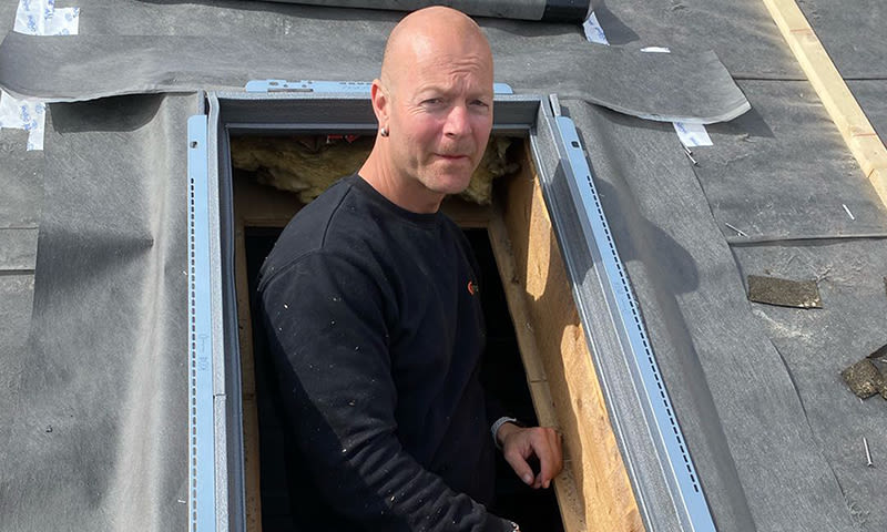 Person i svart genser kikker ut av takvinduåpning med grå metallramme på svart takbelegg.