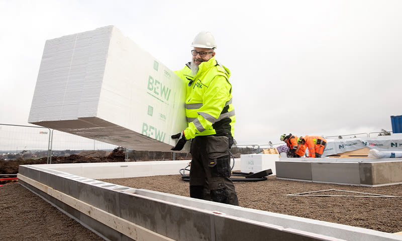Byggearbeider i gul jakke og hvit hjelm løfter stor hvit BEWI-blokk på fundament under grå himmel