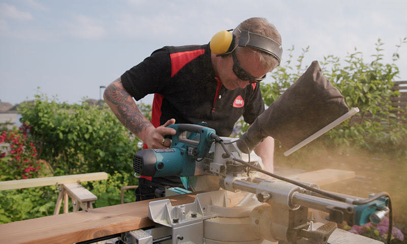 Håndverker i svart-rødt arbeidstøy med gult hørselsvern sager trelist i hage, sagstøv flyr.