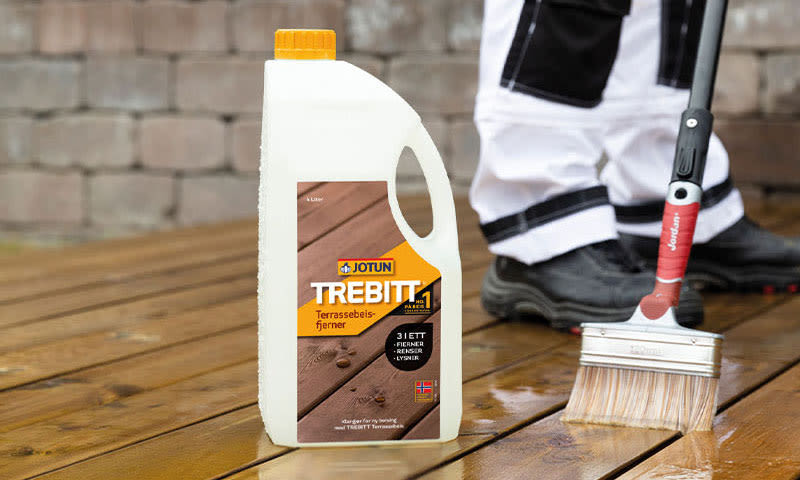 Hvit plastkanne med Jotun Trebitt terrassebeisfjerner på våt treterrasse, malerkost og verneutstyr.