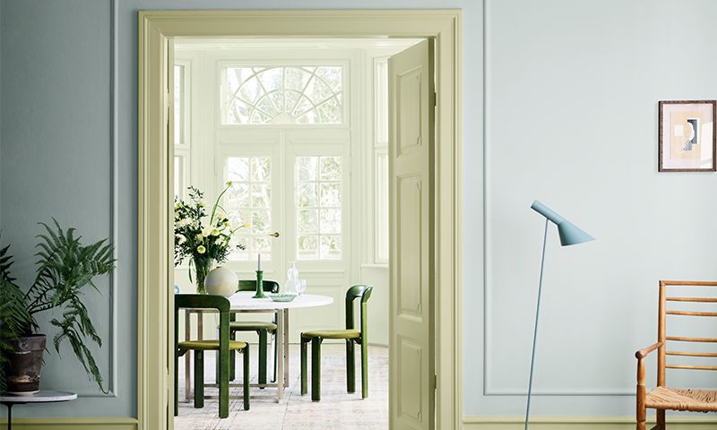 Lyseblå stue med gulnet døråpning mot solfylt spiserom med hvitt bord og grønne stoler