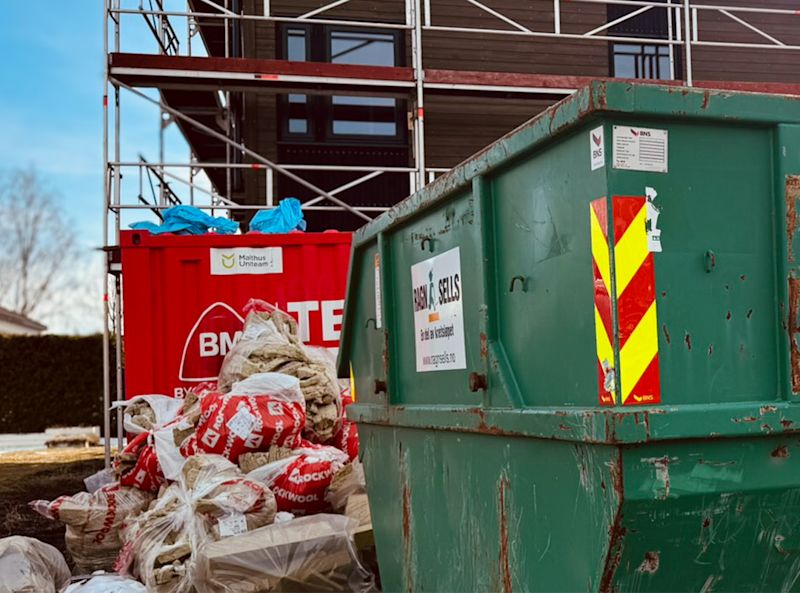 Grønn og rød avfallscontainer på byggeplass med sekker av Rockwool-isolasjon og stillas foran en bygning.
