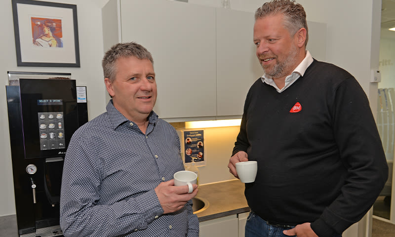 To kolleger ved kaffemaskin i kontorkjøkken, holder hvite kopper ved grå benk.