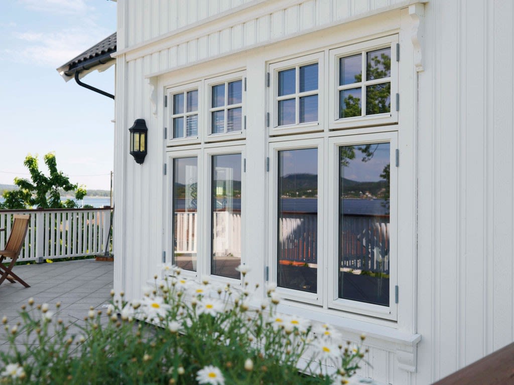 Hvit tømmerhus med store vinduer, prestekrager i forgrunnen, terrasse og sjøutsikt.