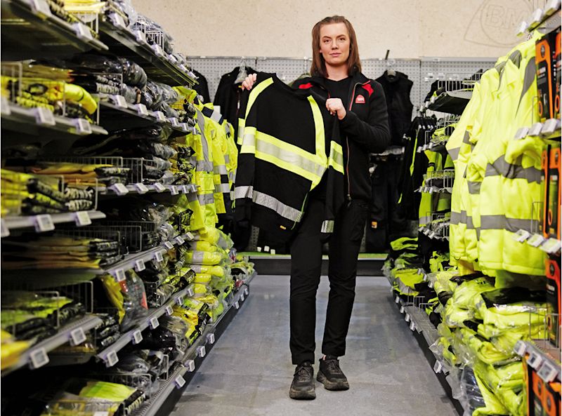 Ansatt holder frem svart jakke med gule reflekser i butikkrekke med gule sikkerhetsklær