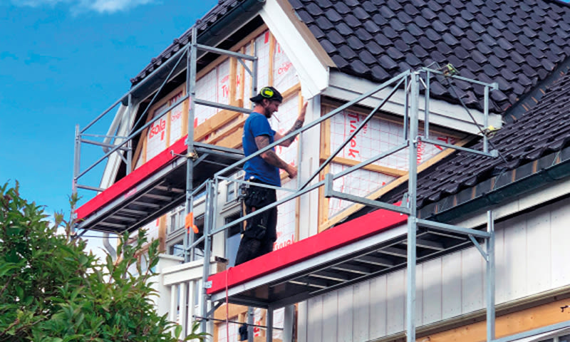 Bygningsarbeider på rødt stillas fester hvite plater på husfasade under blå himmel