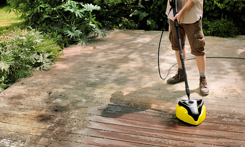 Person med gul og svart høytrykksspyler renser skitten treterrasse i hage.