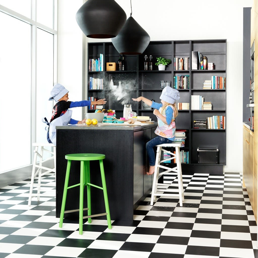 To barn med kokkeluer kaster mel ved sort kjøkkenøy på sort-hvitt gulv med grønn krakk