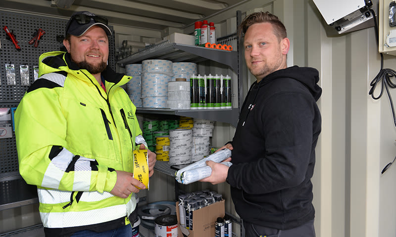 To arbeidere, én i neonjakke og én i svart hettegenser, i container med hyller fulle av tape og lim