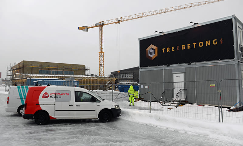 Snødekt byggeplass med gul kran, grå containermoduler med Tre & Betong-logo, hvit/rød varebil