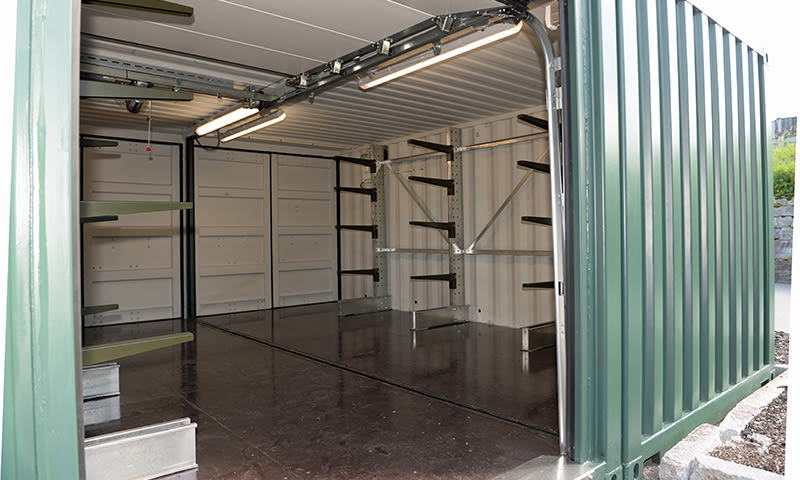 Åpen grønn metallcontainer med tomt, mørkt gulv, hyller og lysrør i taket.