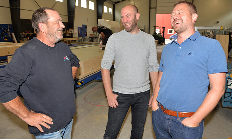 Tre menn i arbeidsklær står i trevarefabrikk med maskiner og trebjelker rundt.