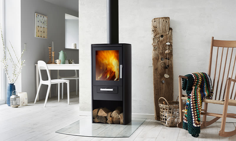 Moderne lyst oppholdsrom med svart peisovn med flammer, ved og gyngestol med fargerikt heklepledd.