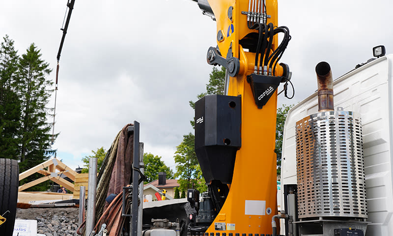 Gul lastebilkran ved treramme på gruslagt tomt under boligbygging, trær i bakgrunnen.