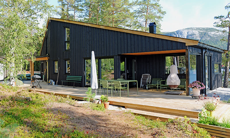 Moderne svart hytte ved innsjø, tømmerterrasse med grønt bord, hvit parasoll, skog og fjell.