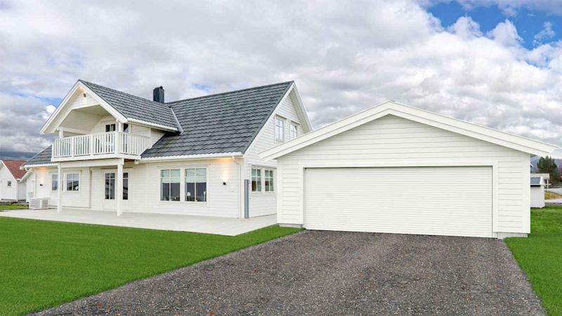 Hvit trevilla med grått tak, dobbelgarasje, grønn plen og skyet himmel.