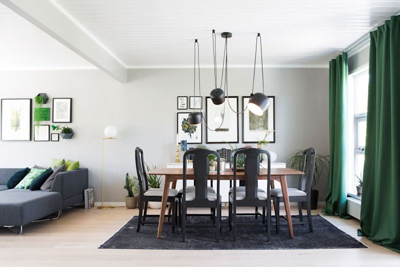 Lyst spise- og stueområde med trebord, svarte stoler, hengende lamper, grønne gardiner og planter.
