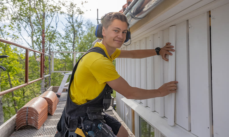 Mann i gul T-skjorte og hørselsvern monterer hvit kledning på stillas utenfor hus