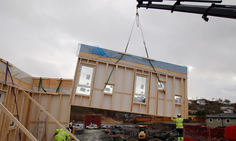 Kran løfter prefabrikkert trevegg med vinduer på byggeplass under overskyet himmel, arbeidere i gult