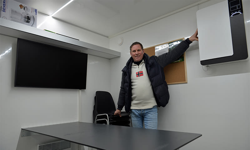 Mann i mørk vest og lys genser med norsk flagg i hvitt møterom med svart TV og ventilasjonsaggregat