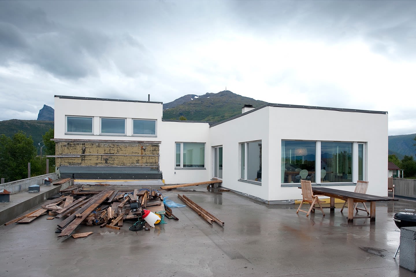 Vått flatt tak med byggeplanker, hvit moderne enebolig, trestoler og fjellandskap bak.