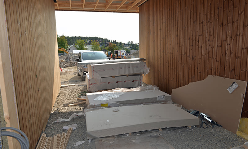 Uferdig garasje med lyse trevegger, stablede gipsplater på grus og hvit varebil i åpningen.