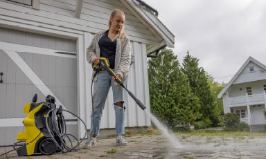 Kvinne bruker gul høytrykksvasker på grå belegningsstein foran hvitt hus og grønne busker.