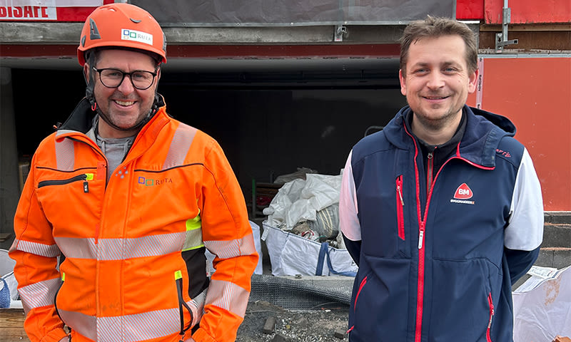 To menn i verneklær ved byggeplass, en i oransje drakt og hjelm, en i blå vest med logo