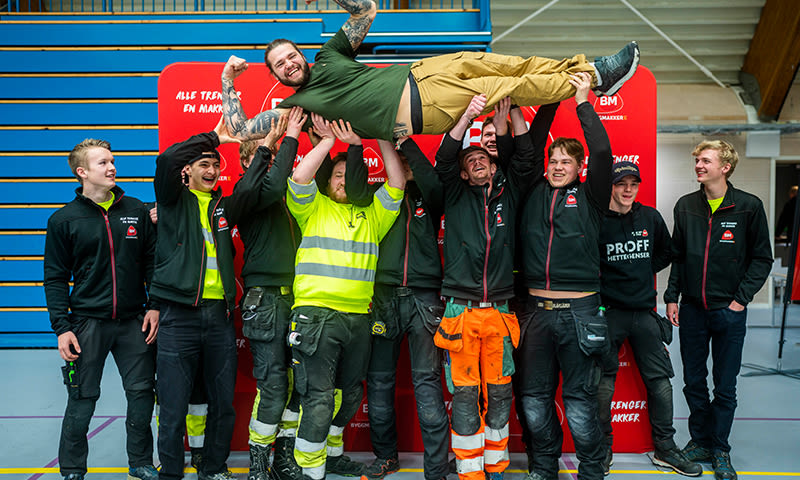 Åtte håndverkere i arbeidsklær og high-vis løfter en kollega over hodet foran rødt banner.
