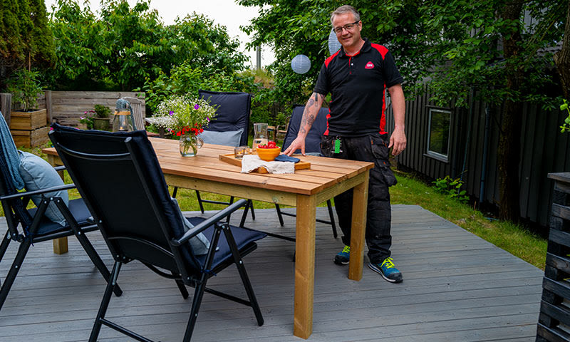 Person i svart-rødt antrekk ved trebord med blomster, skål og stoler på grå terrasse
