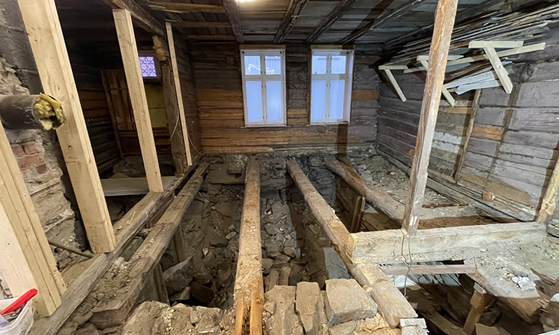 Interiør gammelt tømmerhus under oppussing, avkledd gulv med brune bjelker og murrester, vinduer.