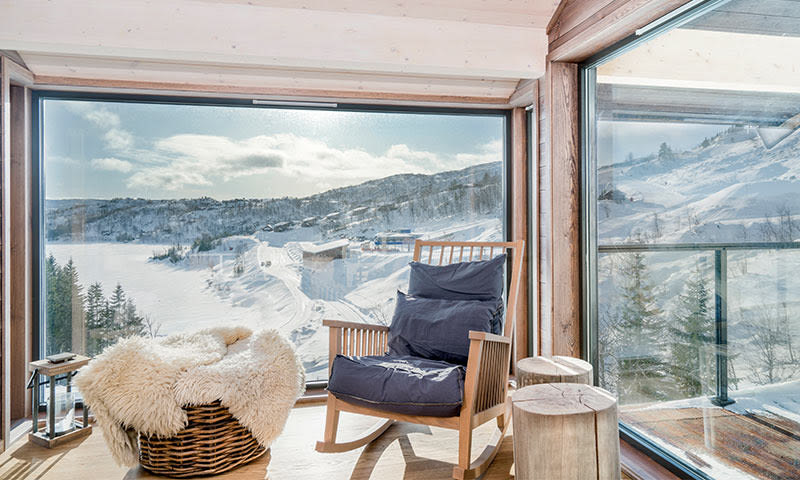 Lyst hytteinteriør med mørk gyngestol, saueskinn og stort vindu mot snødekt fjell