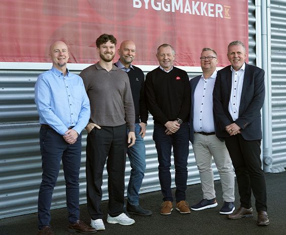 Seks personer står på rekke foran en vegg med Byggmakker-logo, alle smiler mot kameraet.