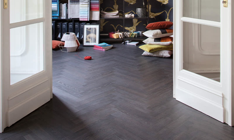 Åpne hvite dører til designstudio med mørk fiskebeinsparkett, fargerike stoffprøver og puter.