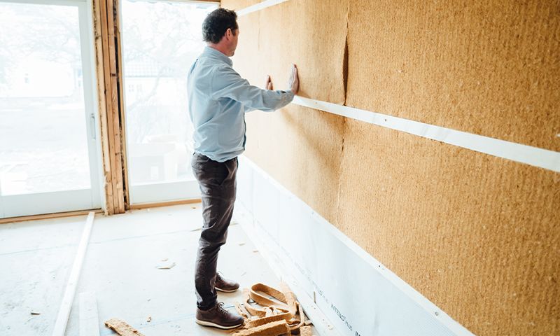 Mann i lyseblå skjorte monterer brun korkplate på vegg i uferdig, lyst oppussingsrom