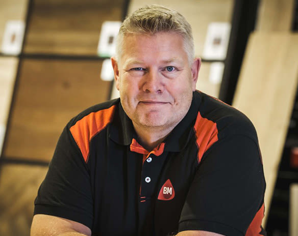 Mann i svart/rød BM-polo foran trepaneler i butikkmiljø.