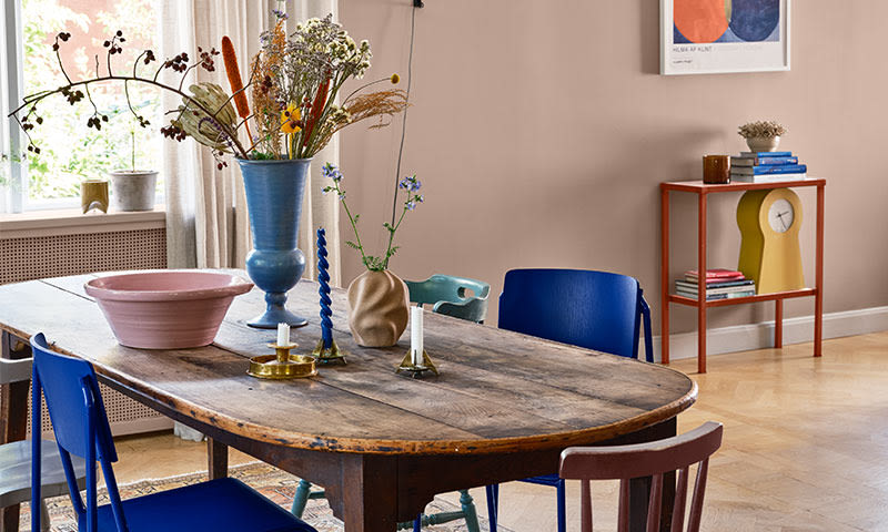 Rustikt trebord med blå stoler, rosa bolle, blomstervaser og lys mot rosa vegg.