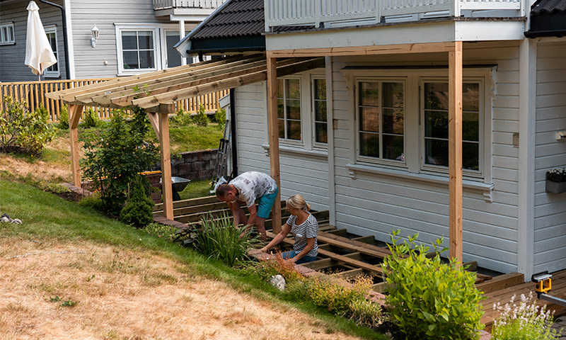 Par bygger treterrasse ved hvit husfasade, gressplen og grønne busker i sol.