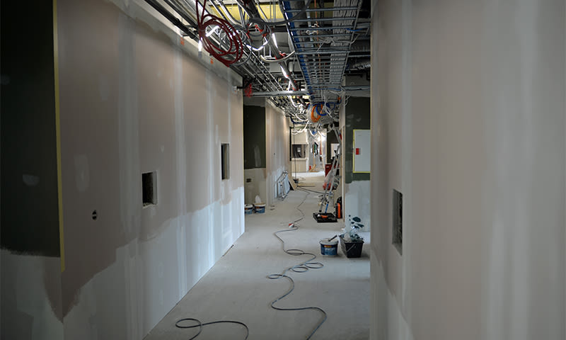 Lang korridor under oppussing med hvite gipsvegger, synlige takk abler og verktøy på gulvet.