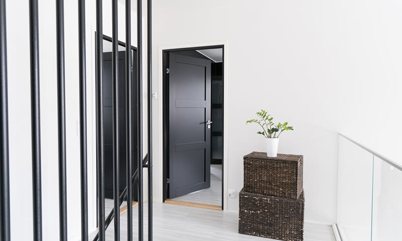 Lyst hvitt gangrom med svarte metallrekkverk, åpen mørk grå dør og kurv med grønn plante.