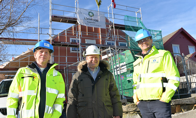 Tre byggearbeidere i gule jakker og hjelmer foran stillasert hus og blå himmel.