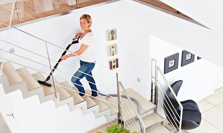 Person i hvit t-skjorte og blå jeans støvsuger hvit trapp med stålrekkverk i lyst hjem.