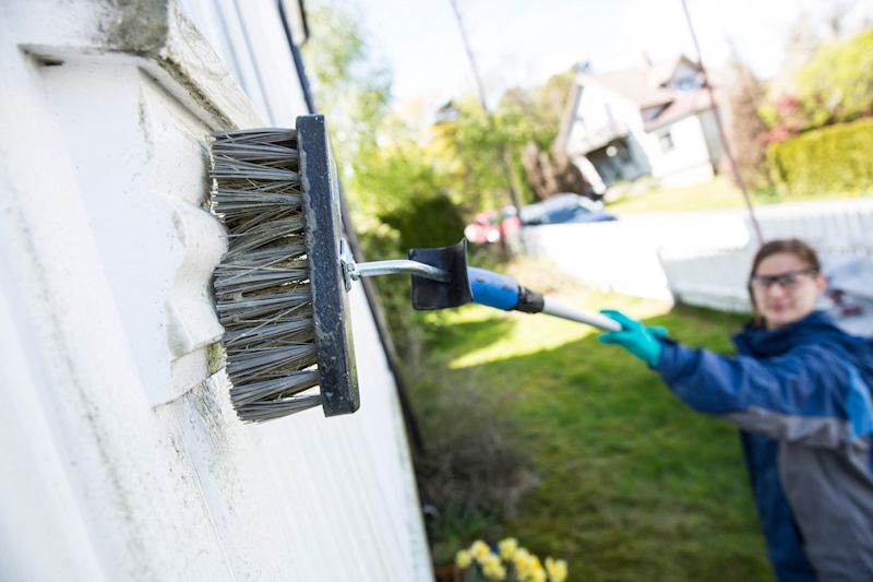 Person i grønne hansker børster hvit husvegg med forlengerskaft i hage